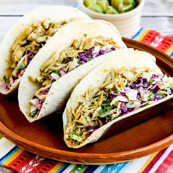 Three Green Chile Chicken Tacos shown in taco holder on plate, with cabbage slaw and limes.