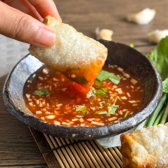 Close-up of fried spring roll dipped into sweet chili garlic sauce.
