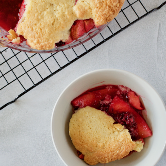 A bowl of Blackberry and Apple Cobbler in a pudding bowl with the larger pie dish behind it on a coo