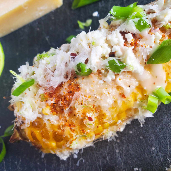 Corn cob buried under toppings on a black cutting board.