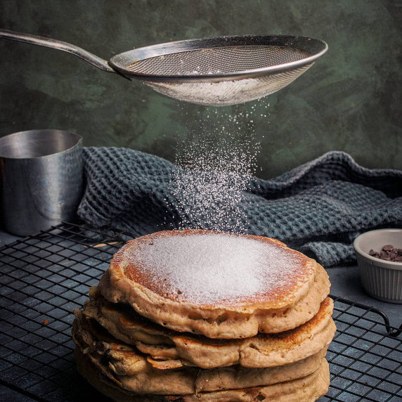 A stack of thick Banana Bread Pancakes sits on a cooling rack while powdered sugar is sprinkled on t