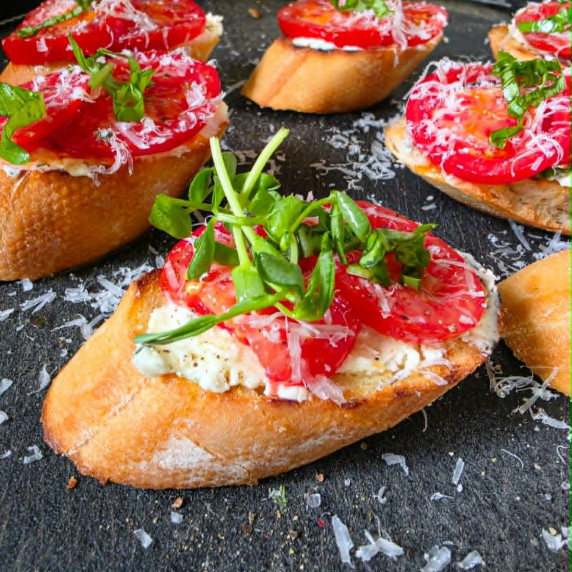 Golden crostini topped with red tomatoes & green herbs on a black board