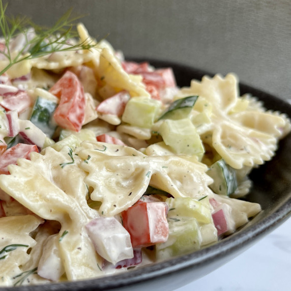 Bow tie pasta, diced cucumber, red pepper and red onion in a black bowl on a marble countertop.