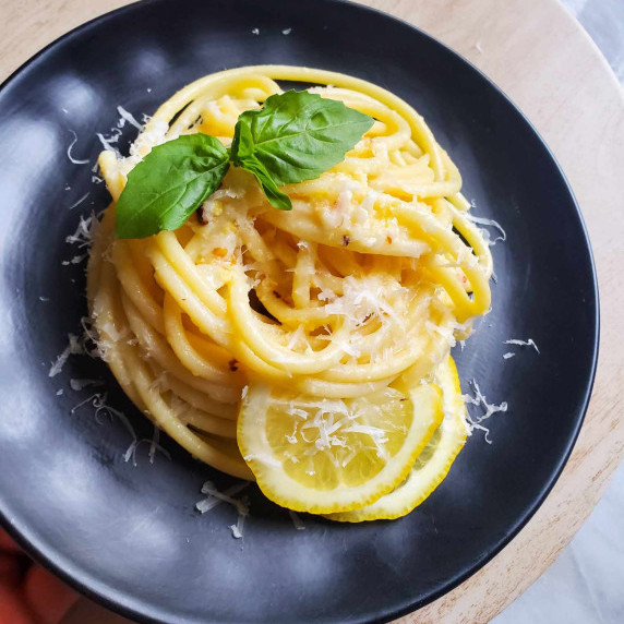 A yellow hued nest of bucatini on a black plate garnished with green, fresh basil