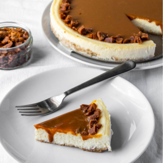 slice of cheesecake with caramel and toffe on a white plate with whole cheesecake behind