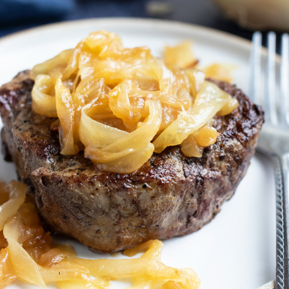 Sautéed Onions for Steak RECIPE served over a steak on a white plate with a fork.