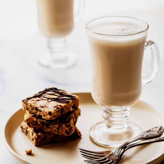 Two chai tea lattes in glass mugs. The front latte is sitting on a creamy white plate with two forks