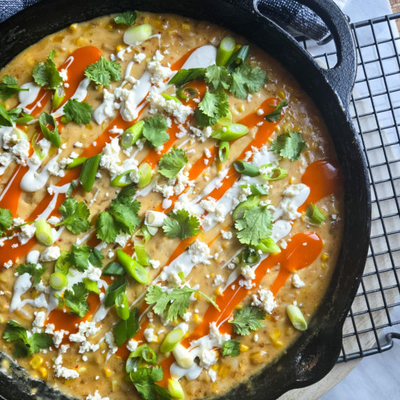 A skillet full of golden corn dip, red buffalo sauce, white dressing, and greens.