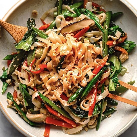 Lo mein noodles, chicken, green onions and red peppers in a white bowl with chopsticks and spoon