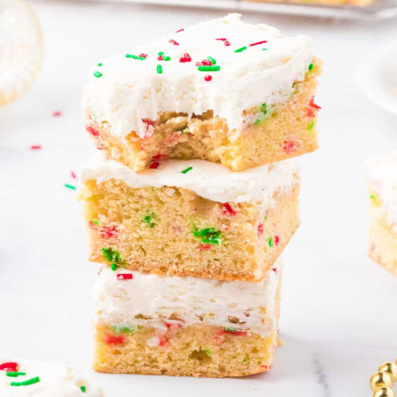 Three frosted sugar cookie bars stacked and covered with sprinkles.  Top bar is missing a bite.