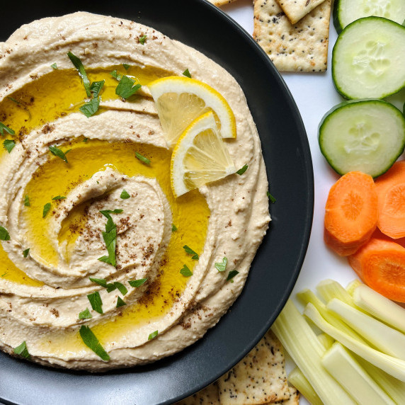 Hummus spread on a black plate drizzled with oil and served with vegetables and crackers.