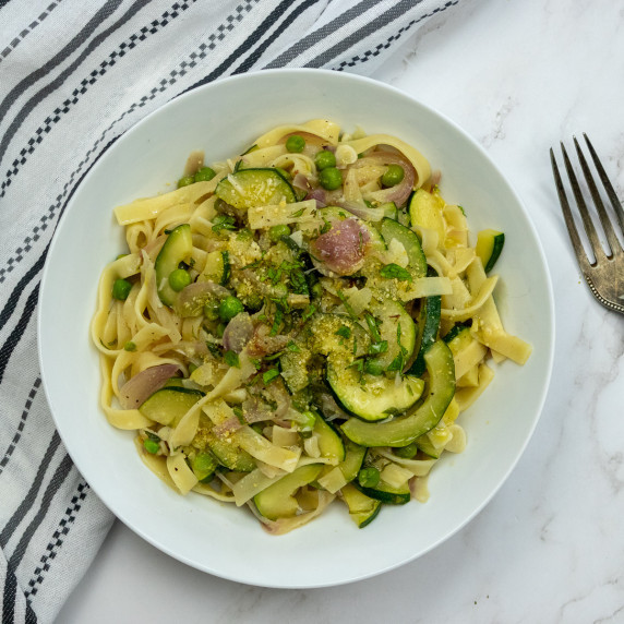 A bowl of vegan zucchini pasta with peas and herbs, served on a white plate with a striped cloth