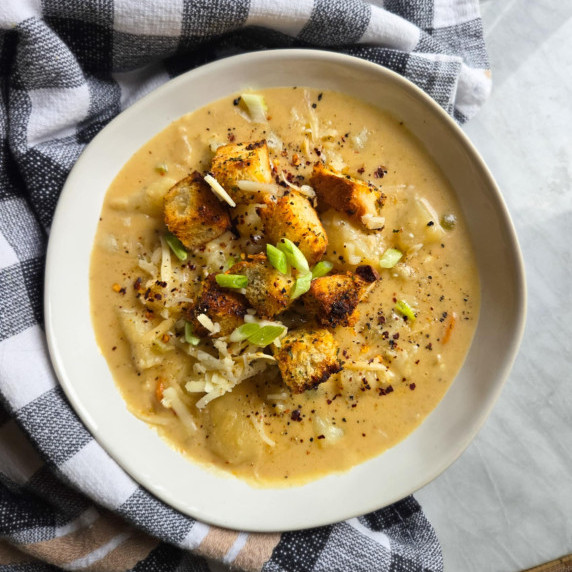 A golden, cheesy potato soup against a checkered towel on white marble.