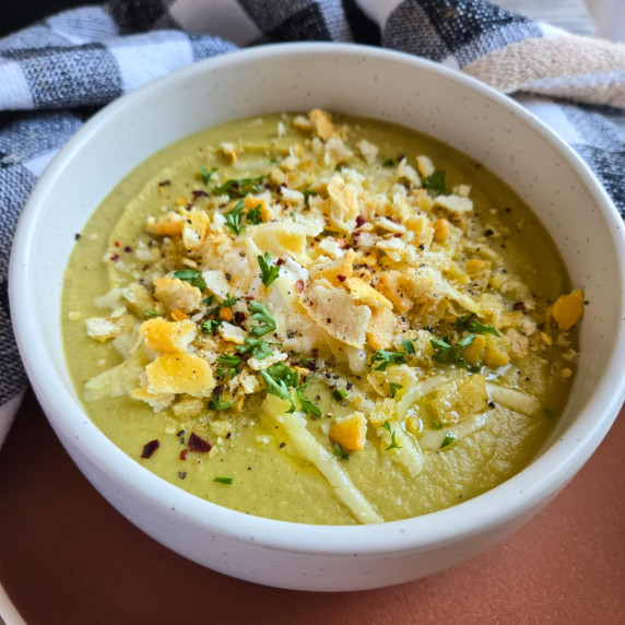 A creamy green soup garnished with crackers & herbs against a checkered towel.