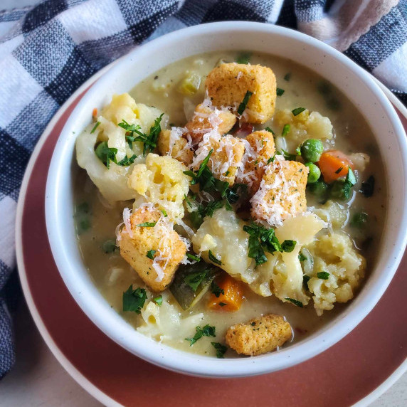 A white bowl filled with chunky veggie stew on a pink plate with a checkered towel behind.