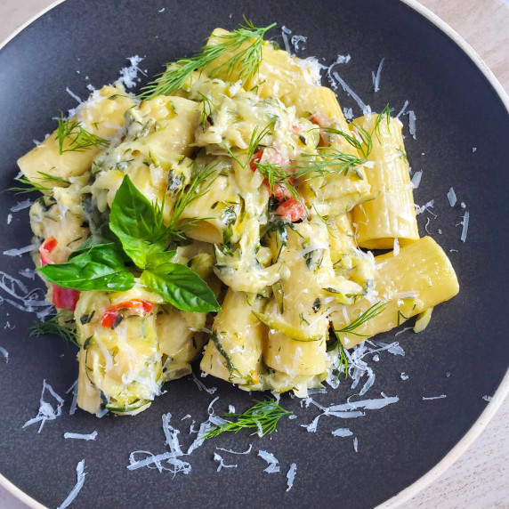 A black plate of rigatoni with fresh green herbs and white shreds of grana padano.