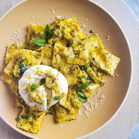 Ravioli dressed in bright pesto with creamy white burrata on a beige plate.