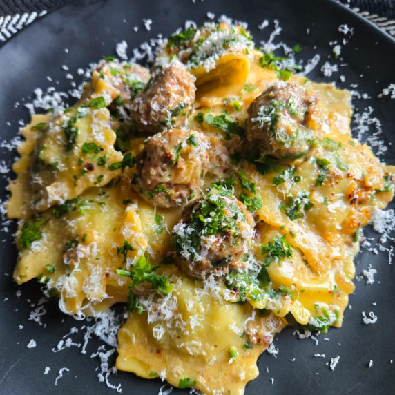 Golden ravioli, golden brown meatballs, and pops of green parsley on a black plate.