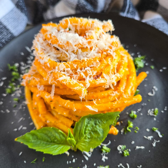 A nest of bucatini in an orange hued sauce with fresh, green herbs on a black plate.