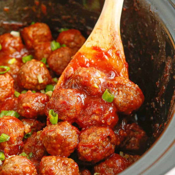 Meatballs close up in a red sauce being lifted from a slow cooker by a spoon.