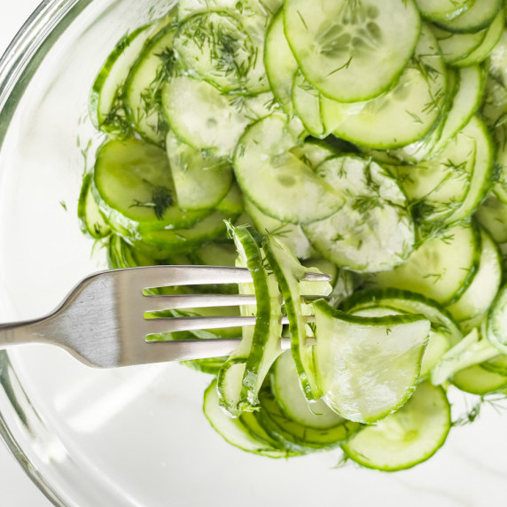 A glass bowl filled with very thinly sliced cucumbers. A fork is picking up a few slices on the left