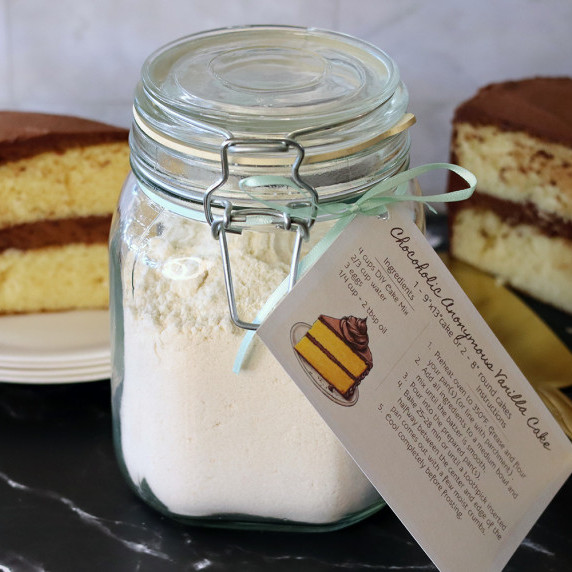 Jar of DIY Vanilla Cake Mix with attached recipe in front of a yellow cake with chocolate frosting.