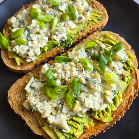 Golden toast topped with green avocado and creamy egg salad on a black plate.
