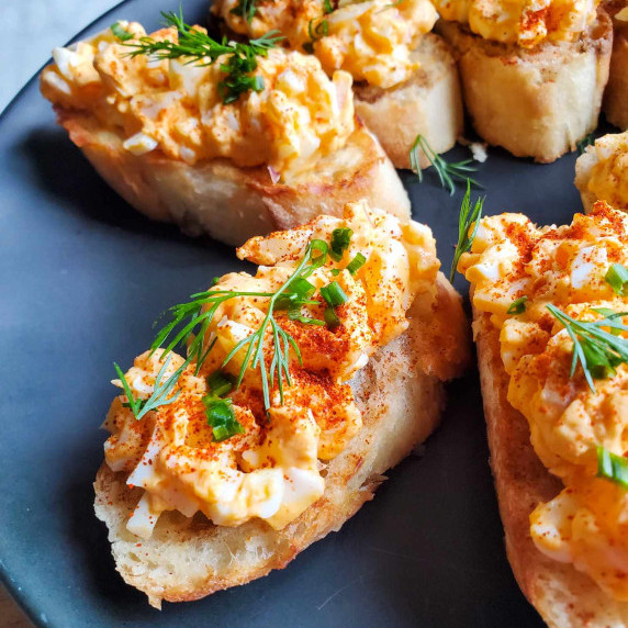 Golden egg salad on top of crispy baguettes on a black plate with pops of green