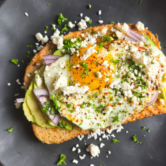 A golden piece of toast topped with green avo, fried egg, pops of white feta and green herbs.