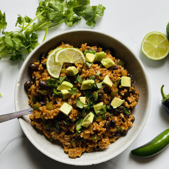 Bowl of enchilada rice garnished with diced avocado, lime wedges and cilantro on a marble board.