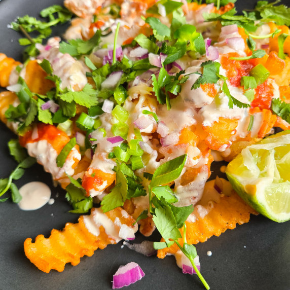 A black plate of golden crinkle fries topped with a variety of colourful garnishes.