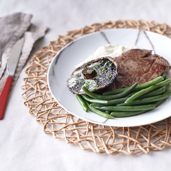 Garlic-covered mushroom on grilled rump steak with cauliflower mash and green beans on a white plate