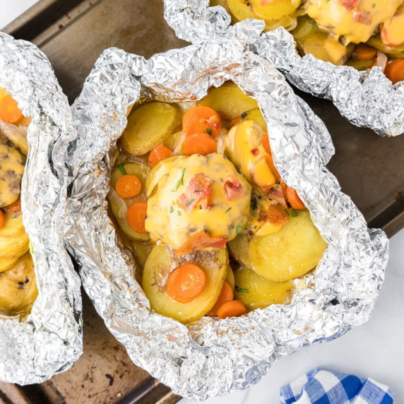 Close up 3 foil packets from above full of sliced potatoes, carrots, onions and burgers with bacon.