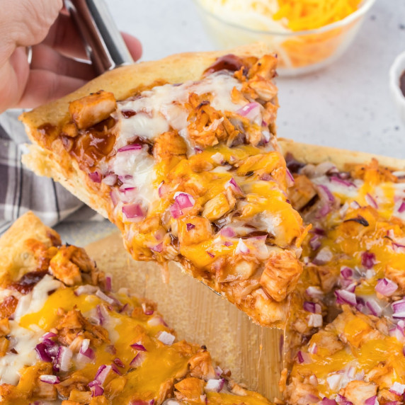 A slice of BBQ chicken pizza on a wooden cutting board being lifted with a spatula.