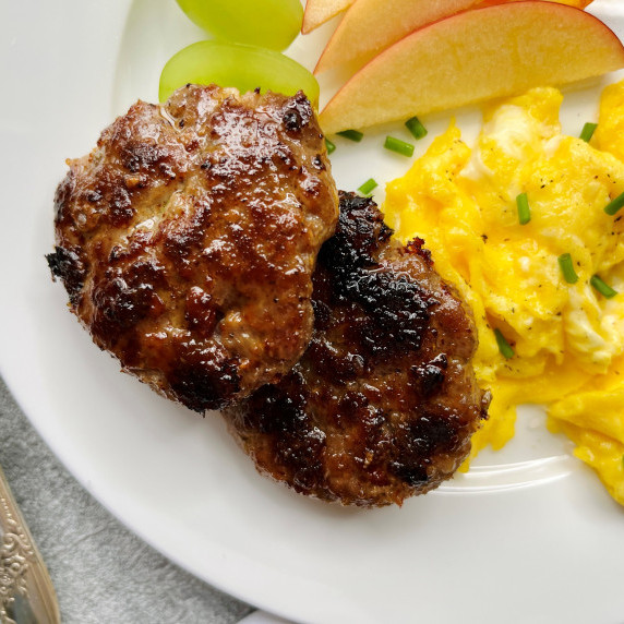 Two sausage patties on a white plate with scrambled eggs and fresh fruit.