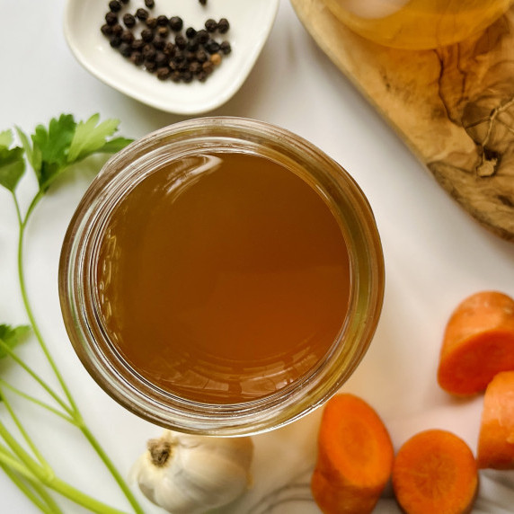 Jar of vegetable broth next to carrots, garlic, parsley and peppercorns on a white counter.