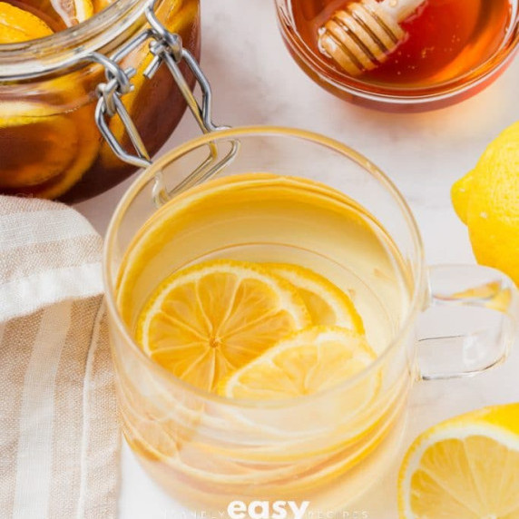 Cup of water with honeyed lemon slices, with lemon fruits and a bowl of honey on the side