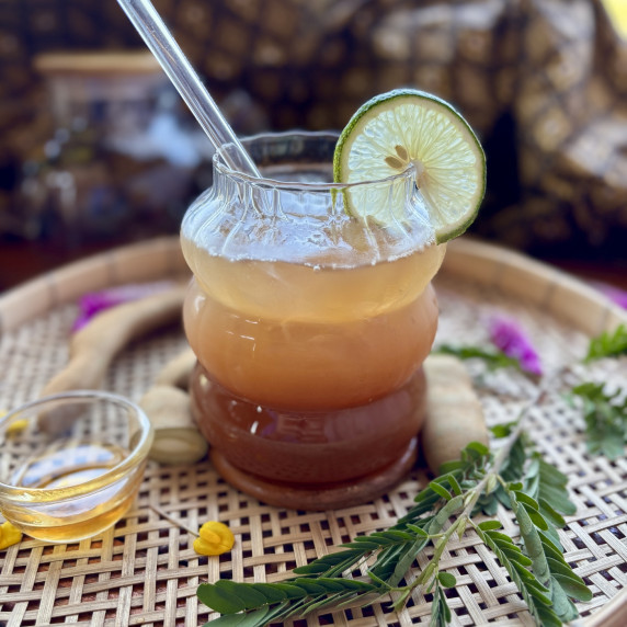 Tamarind iced tea in a glass, with tamarind pods and leaves, and a cup of honey surrounding it.