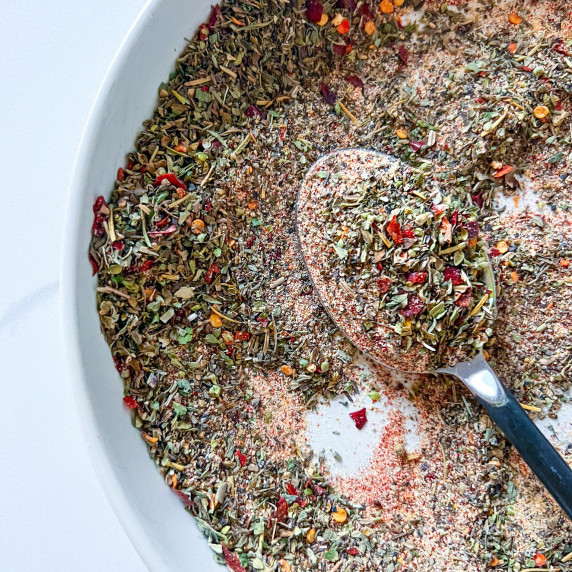Pizza seasoning in a white bowl with a spoon. You can see bits of the chili flake and herbs.