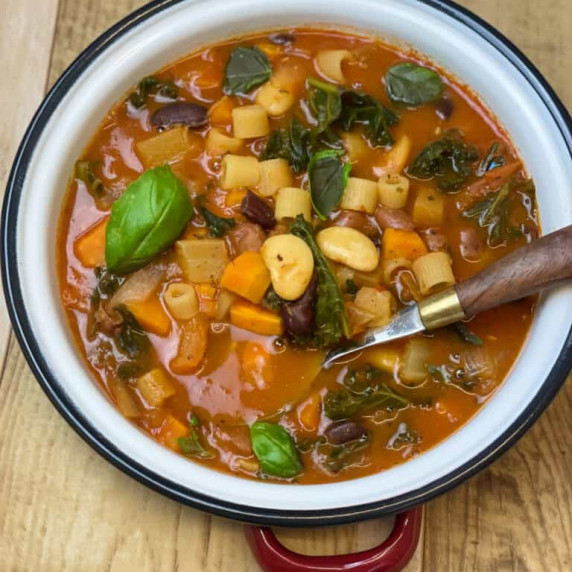 A white bowl with black rim full of bean soup with basil garnish, wooden background.