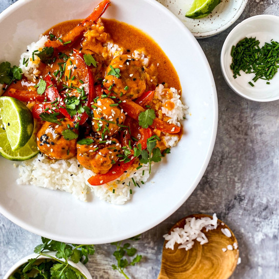 A white plate with chicken meatballs in a red curry peanut sauce, over white rice on a grey surface.