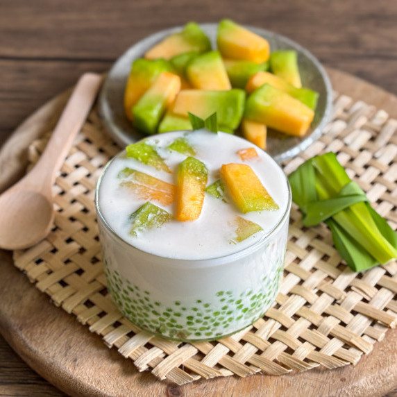 Thai coconut melon sago served in a glass cup, with ripe mango in the background.