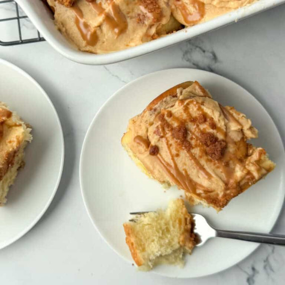 Overhead photo of Biscoff Cinnamon Roll on a plate with a bite taken out in front of a pan of rolls
