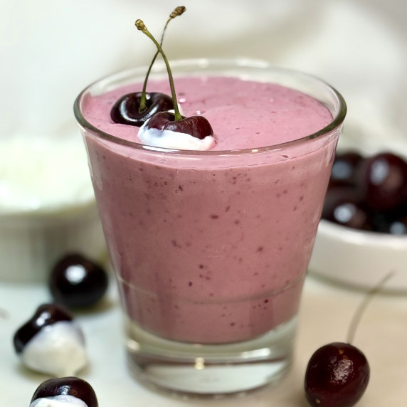 A glass of pink cherry vanilla smoothie is seen on a white counter, surrounded by fresh cherries.