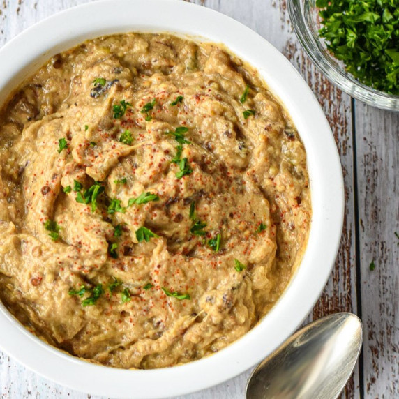 instant pot baba ganoush in a white bowl next to a spoon and bowl of chopped parsley