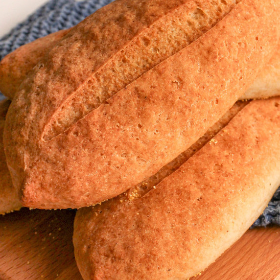 Gluten free Italian sub rolls, stacked on wood cutting board with blue kitchen towel