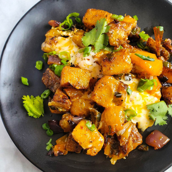 A seasoned sauté of potatoes and veg on a black plate against a marble background. 