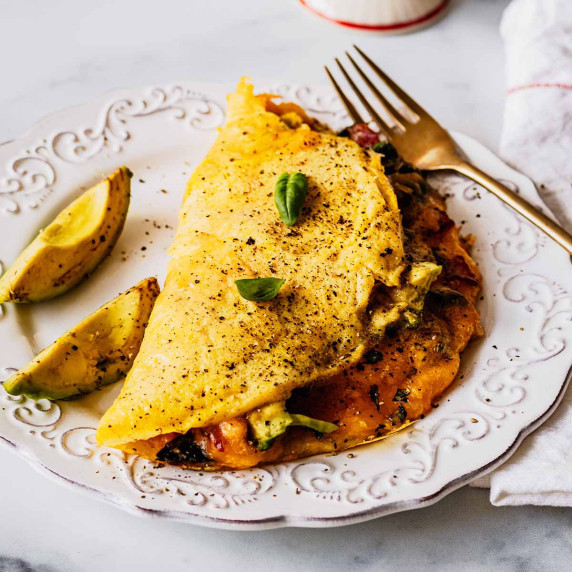 Keto omelette topped with basil leaves on a white plate with sliced avocado on the side