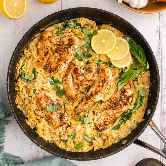 Fry pan filled with golden yellow orzo and lemon chicken, topped with basil leaves as garnish.
