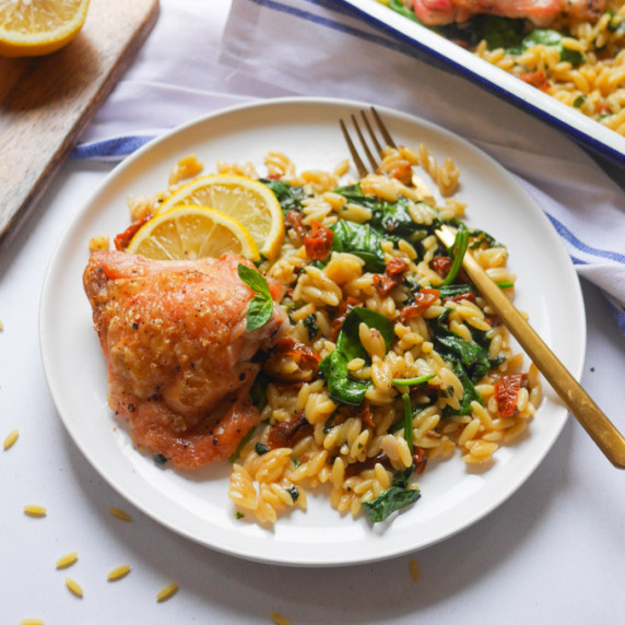 A roast chicken thigh on a plate with orzo, sundried tomato, spinach, lemon and basil.